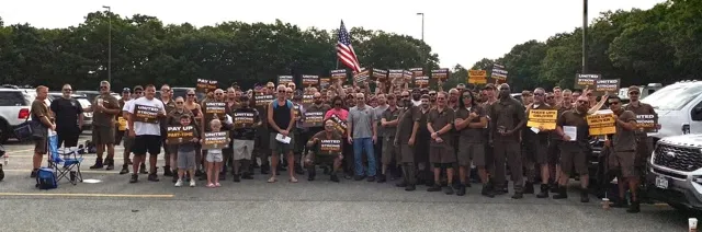 Around a hundred UPS workers in brown uniforms stand together across a parking lot behind an American flag.