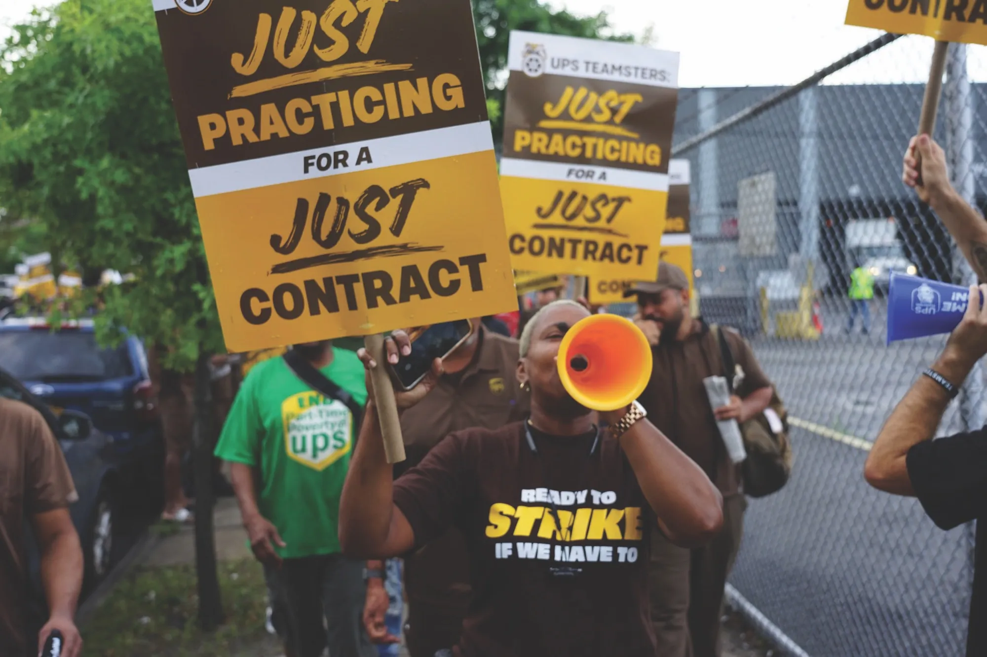 A Local 804 member leads a chant through a bullhorn on a picket line, beneath signs reading “Just practicing for a just contract”.