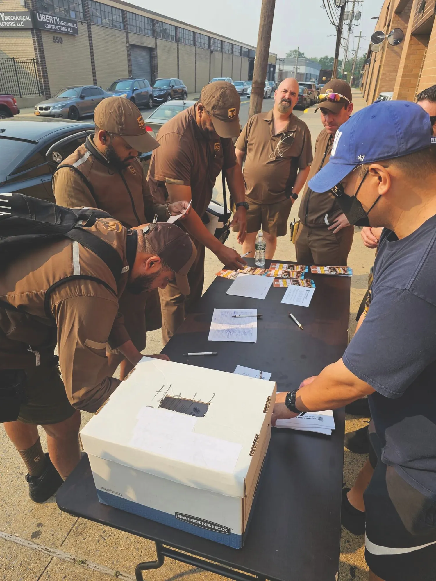 UPS drivers in brown uniforms crowd around a folding table to cast strike-vote ballots on a Queens street.